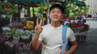Şehrin hareketli caddesinde renkli çiçekçinin önünde barış işareti yapan genç adam dışarıda mutluluk ve pozitiflik yayıyor.