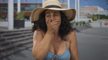 Orta yaşlı, bikini giyen ve güneş şapkalı İspanyol bir kadın güneşli bir sokakta açık havada neşeli ve şaşırtıcı bir ifade sergiliyor..