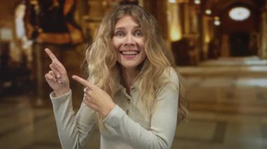 Kadın elini kaldırır ve salona doğru işaret eder. Bu sırada sarışın saçı, müze lobisinde karmaşık mimarisi olan heyecanlı bir genç figürü çerçeveler..