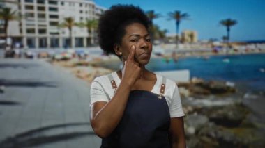 Önlük giymiş bir kadın, parmakları yanaklarında, canlı bir plaj zeminine, palmiye ağaçları ve mavi gökyüzüne karşı düşünceli bir ifade sergiliyor..