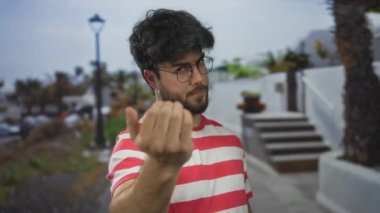 Genç, sakallı, İspanyol bir adam güneşli bir günde, arka planda palmiye ağaçları ve beyaz merdivenlerle gelmesi için el hareketi yapıyor. Rahat bir ortam izlenimi veriyor..