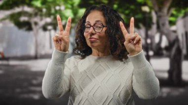 Beyaz kazaklı, kıvırcık saçlı bir kadın şehir sokaklarında jestler yapıyor. Arka planda ağaçlar, açık havada gülümserken neşe ve güveni ifade ediyor..