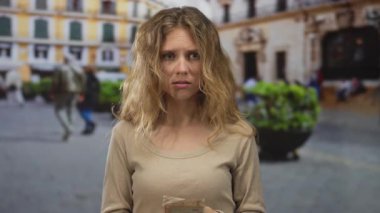 Bir plazada nakit para sayarken elinde banknot tutan bir kadın, para sıkıntısı ve endişeli bir sokak ifadesiyle sarışın bir genç olarak ortaya çıkıyor..