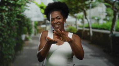 Parkta beyaz atlet giyen bir kadın, elleri ağaçların altında gülümseyen bir jest yapıyor..