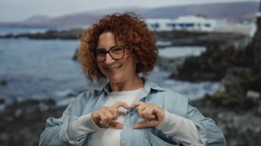 Middle-aged hispanic woman smiling at seaside beach making heart gesture against picturesque sea background