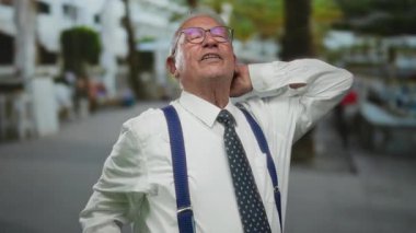 Takım elbiseli son sınıf öğrencisi, arka planda ağaçlar olan canlı bir şehir ortamında gözlük ve pantolon askısı takıp, bir sokakta kendinden emin bir şekilde duruyor..