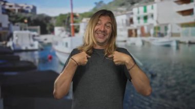 Koyu gri tişörtlü, uzun sarı saçlı bir adam her iki işaret parmağıyla binanın önünü işaret ediyor..