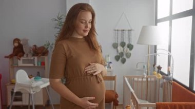 Hamile kadın, pencerenin yanındaki bir binada, karnına hafifçe dokunur ve gün ışığında lamba yakar..