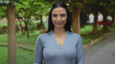 Genç İspanyol bir kadın güneşli bir parkta ağaçlarla ve arka planda bir patikayla bekliyor. Açık havada bir beklenti ve beklenti anı yakalıyor..