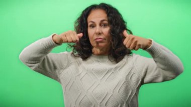 Kıvırcık saçlı bir kadın yeşil arka planda yukarı aşağı başparmak gösteriyor. Stüdyo ortamında karışık duyguları canlandırıyor..