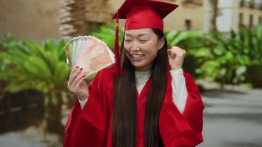 Elinde Filipinler Peso banknotları olan kırmızı mezuniyet elbisesi giymiş bir kadın arka planda yemyeşil bir yeşillikle neşe içinde dışarı çıkıyor..