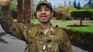 Kamuflaj üniformalı bir adam ve sokakta palmiye ağaçlarıyla el sallayan künye kolyesi; görev ve gurur..