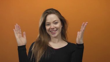 Genç gülümseyen beyaz kadın, stüdyoda iki avucunu da turuncu duvarla kafasının yanına kaldırıyor; neşeli açıklık.