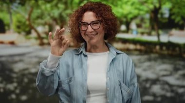Parkta gülümseyen bir kadın, gözlük takan ve günlük kıyafetler giyen, parlak bir ifadeyle baş parmak işareti yapan, güneşli bir günde yeşil ağaçlarla çevrili..
