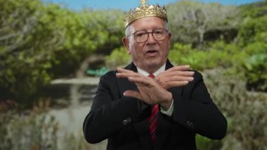 Takım elbiseli kıdemli bir adam taç takıyor. Parkta parmakları yeşil ağaçlarla, manzaralı arka planda, iş kıyafetleri giyip otoriteyi ve ihtişamı ima ediyor.