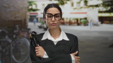 Siyah cüppeli ve büyük gözlüklü yargıç, şehir caddesinde tokmağı kavrayıp, otoriteyi ve profesyonelliği somutlaştıran kollarıyla kendinden emin bir şekilde duruyor..