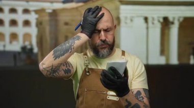 Sakallı ve dövmeli, şehir ortamında eldiven ve tulum giymiş, açık havada notlara odaklanmış düşünceli bir yüz ifadesi olan, konsantre olmuş bir adam..