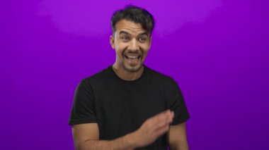 Genç İspanyol bir adam, izole edilmiş mor arka planda fısıldıyor. Siyah gömlekli, neşeli bir ifadeyle neşe ve gizlilik gösteriyor.