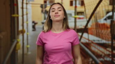 Pembe tişörtlü genç bir kadın, inşaat alanında, etrafı inşaat iskelesi ve şehir dekoruyla çevrili bir şekilde, bağlılık ve samimiyetini ifade ederek yemin jesti yapıyor..