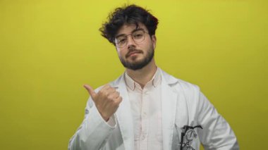 Beyaz önlüklü, sakallı ve gözlüklü genç İspanyol adam, sağlıklı bir ortamda onay ve pozitiflik öneren canlı sarı arka plana karşı emin adımlarla işaret ediyor..