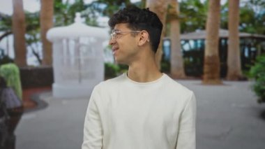 Gözlüklü bir adam ormanda açık palmiyelerle omuz silker yapraklı ağaçlar ve beyaz çeşme ile çerçevelenir; belirsizlik kafa karışıklığı.