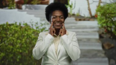 Beyaz ceketli kadın, yeşil ve taş basamaklı güneşli parkta konuşma egzersizleri yapıyor. Dışarıdaki iletişimi ve rahatlamayı ön plana çıkarıyor..