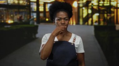 Önlüklü kadın arka planında bulanık ışıklarla şehir caddesinde jest yapıyor. Gençlik enerjisini ve kentsel enerjiyi ön plana çıkarıyor..