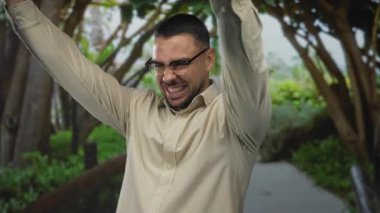 Gözlüklü İspanyol genç adam yeşil parkta sıkılmış yumruklarını kaldırıyor ve sırıtıyor. Zafer sevinç kutlamaları..