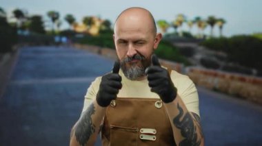 Önlüklü ve eldivenli kel bir adam açık gökyüzünün altında görünen dövmeleri olan bir şehir sokağını işaret ediyor..