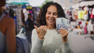 Elinde Tayvan banknotlarıyla gülümseyen bir kadın açık hava hediyelik eşya dükkanında el kol gezerken canlı bir pazar ortamında alışveriş yapmanın ve seyahat etmenin heyecanını somutlaştırıyor..