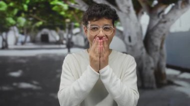 Gözlüklü ve beyaz kazaklı bir adam iki eliyle şehrin caddesinde ağaç gövdesi ve bina duvarı ile ağzını kapatıyor..