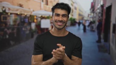 Siyah gömlek giyen ve gümüş bilezik takan adam, güneşli bir öğleden sonra sokakta gülümserken el çırpıyor;.