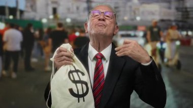Takım elbiseli kıdemli adam elinde dolar sembolü olan para çantası tutuyor ve kalabalık bir sokakta iyi bir jest yapıyor..