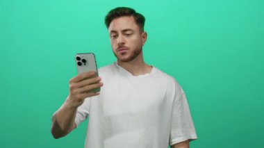 Stüdyo ortamında izole edilmiş yeşil arka planda akıllı telefon tutan sakallı genç adam gündelik kıyafetlerle modern teknoloji ve iletişim temasını gösteriyor..