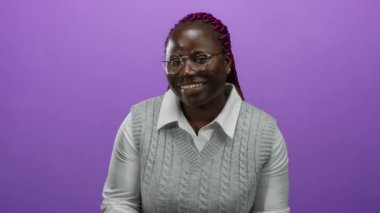 Gözlüklü ve gri süveterli, mor arka planda elleri havada gülümseyen Afrikalı Amerikalı bir kadın..