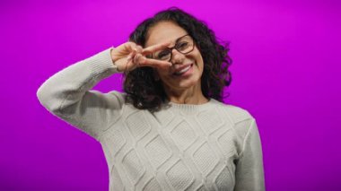 Kıvırcık saçlı, gülümseyen ve barış simgesiyle poz veren bir kadın canlı mor arka planda gözlük takıyor ve örgü süveter örüyor neşeli ve ulaşılabilir bir ortam yaratıyor.