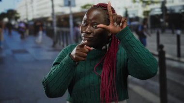 Örgülü bir kadın ezik jestler yapıyor ve şehir caddesinde canlı bir şehir ortamında kameraya bakıyor..