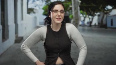 Beton binaları ve güneşli havası olan, gözlüklü ve günlük giysileri olan, sakin şehir atmosferiyle çevrili bir caddede güvenle duran bir kadın..