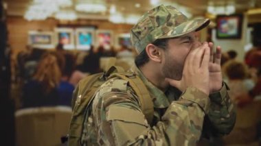 Kamuflaj üniformalı İspanyol adam elleri ağızda bulanık seyirci ve renkli çerçeveli resimlerle bina içinde bağırıyor; görev disiplini odaklanma kararlılığı.