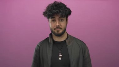 Sakallı genç İspanyol adam canlı pembe bir arka plana el çırpıyor, sıradan moda ve çekici bir ifade sergiliyor..