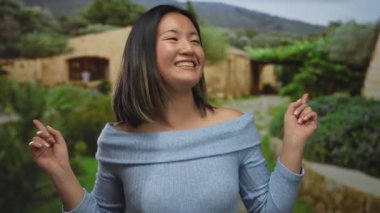 Arka planda taştan yapılar ve neşe ve enerji saçan, yemyeşil bir parkta dikilen ve farklı yönlere gülümseyen bir kadın..