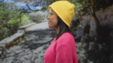 Pembe kazaklı, sarı şapkalı, gülümseyen ve parmaklarını açık havada güneş ışığının altındaki yeşil bir parkta, mutluluk ve gençlik enerjisini somutlaştıran bir kadın..