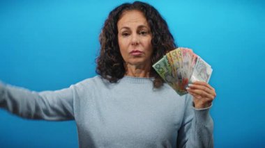 Avustralya doları tutan orta yaşlı bir kadın, mavi izole edilmiş bir arka plana selam veriyor, ışık ayarında para gösteriyor..