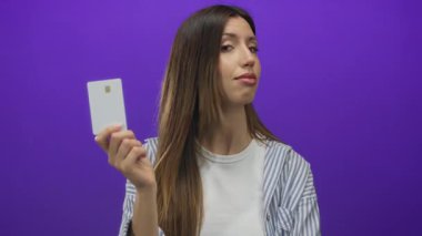 Genç bir kadın mor stüdyo duvarına karşı ciddi bir ifadeyle ve kararlı bir bakışla kredi kartı tutuyor..