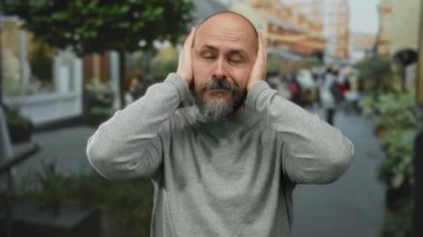 Sakallı kel adam şehir merkezinde kulaklarını kapatıyor. Gri süveter giyiyor. Şehir ortamında gürültü ya da konsantrasyon öneriyor..