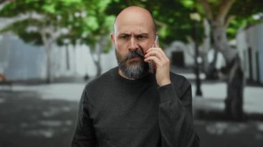 Şehir merkezinde sakallı bir adam telefonda konuşuyor. Yüzünde ciddi bir ifade var. Görünür dövmesi var..