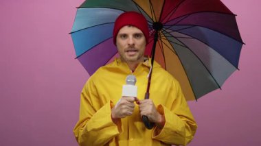 Sarı yağmurluklu adam mikrofon tutuyor. Pembe arka plana karşı çok renkli şemsiye altında duruyor.
