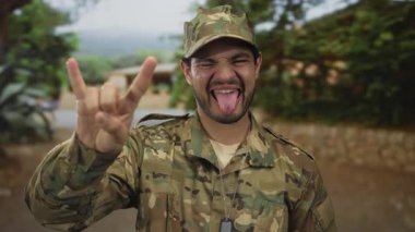 Kamuflaj üniformalı, künye takan bir adam taştan bina cephesinin yanında eski kasaba sokaklarında boynuz hareketi yapıyor..