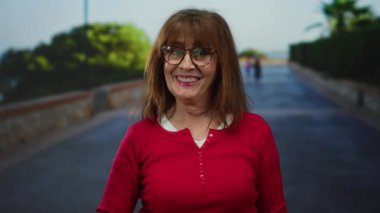 Kırmızı kazaklı kıdemli İspanyol kadın, başparmağıyla şehir dışındaki bir sokakta gülümsüyor ve kentsel bir çevrede neşe ve pozitiflik hissi veriyor..