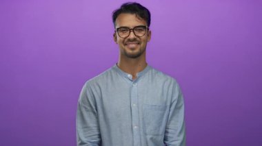 Genç İspanyol bir adam gülümsüyor ve parlak mor bir arka plana el sallıyor. Gözlük takıyor ve mavi bir gömlek giyiyor. Stüdyoda arkadaşlığı ve sıcaklığı somutlaştırıyor..